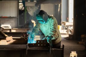 A welder wearing protective gear works in a workshop, welding metal with visible sparks.
